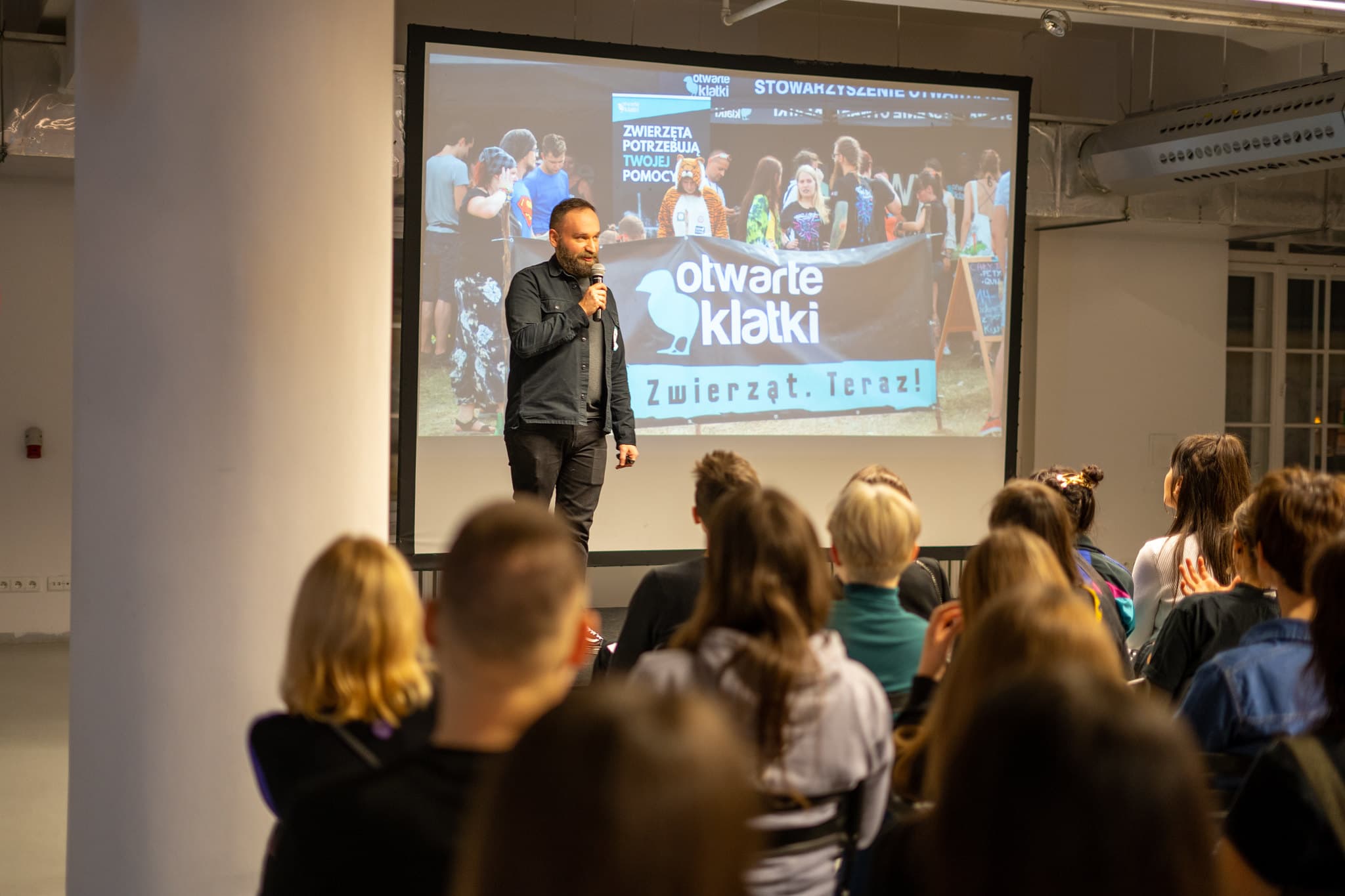 Mężczyzna na scenie, prowadzący wykład, w tle zdjęcie z banerem Otwartych Klatek