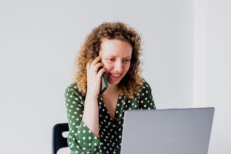 Uśmiechnięta kobieta w zielonej koszuli w białe kropki rozmawia przez telefon, siedząc przed laptopem.